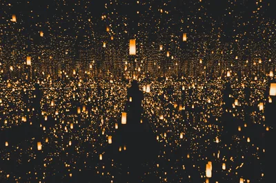 Countless warm lanterns float in layered darkness, doubled by mirror reflections, with a dark silhouetted shape rising through the middle of the frame.