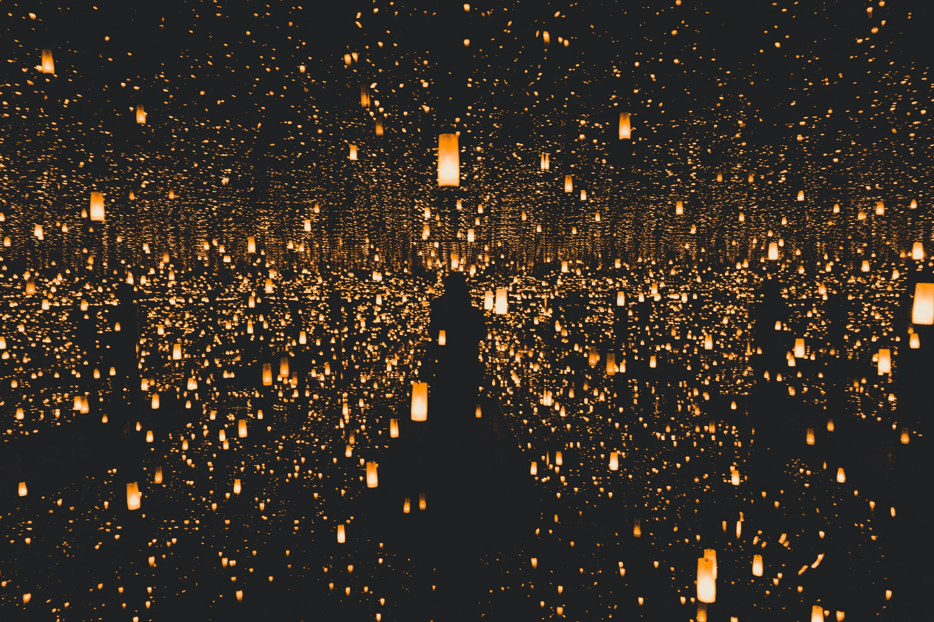 Countless warm lanterns float in layered darkness, doubled by mirror reflections, with a dark silhouetted shape rising through the middle of the frame.