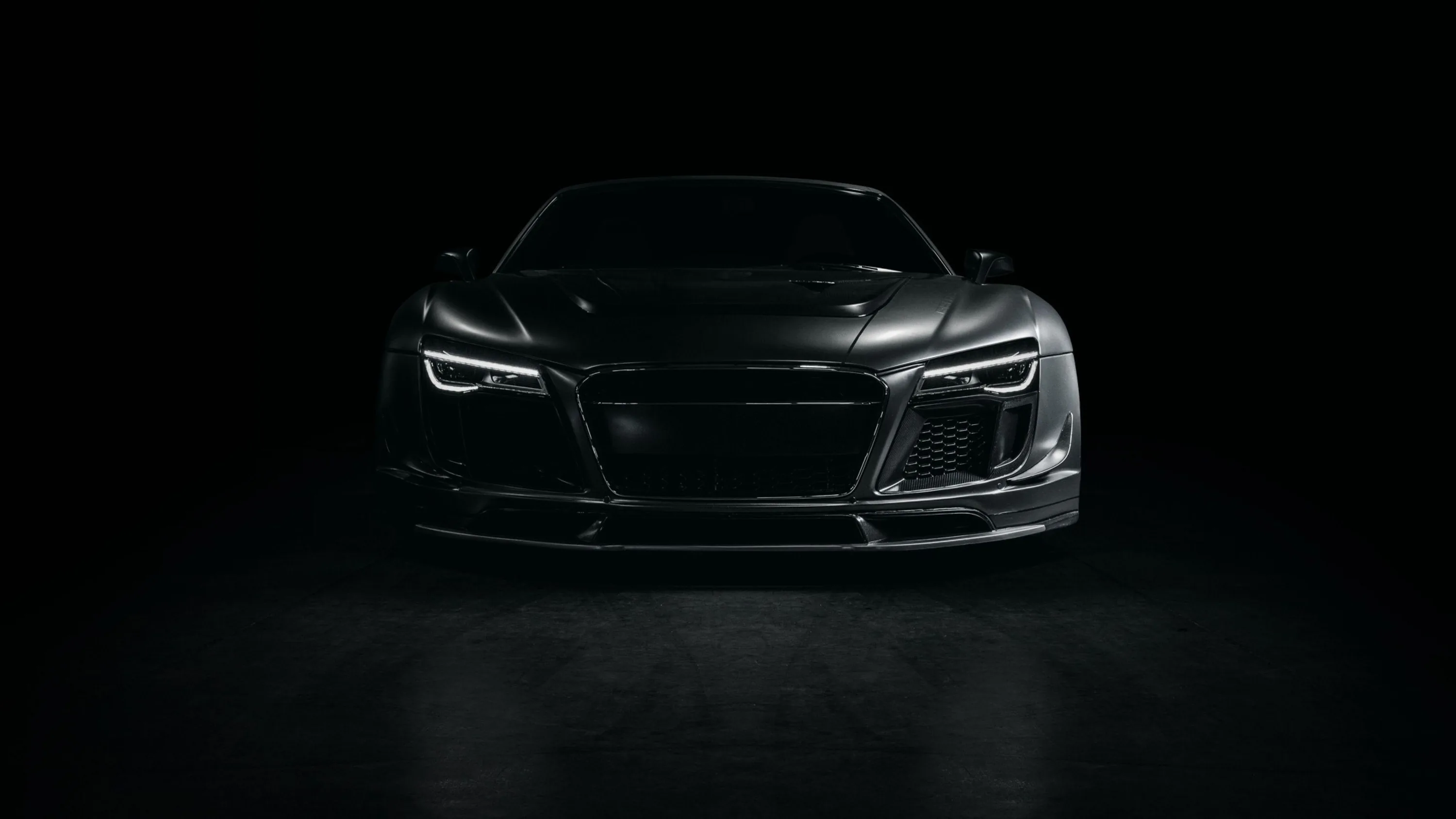A car faces the camera head-on in a black studio, its headlights and grille the only clearly lit parts.