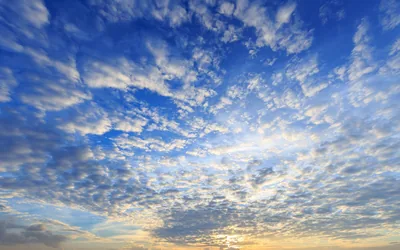 Small dappled clouds cover a blue sky, catching gold from a low sun just below centre frame, where the light is brightest.