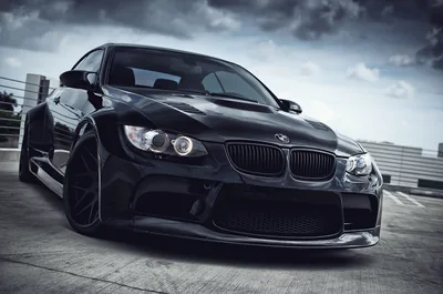 Low front three-quarter of an E92 in black on wide wheels, storm cloud overhead and a chain-link fence running off to the left.