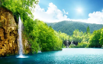 Water sheets down a rust-stained cliff at left while the sun flares in the top corner, throwing a bright track across the turquoise lake.