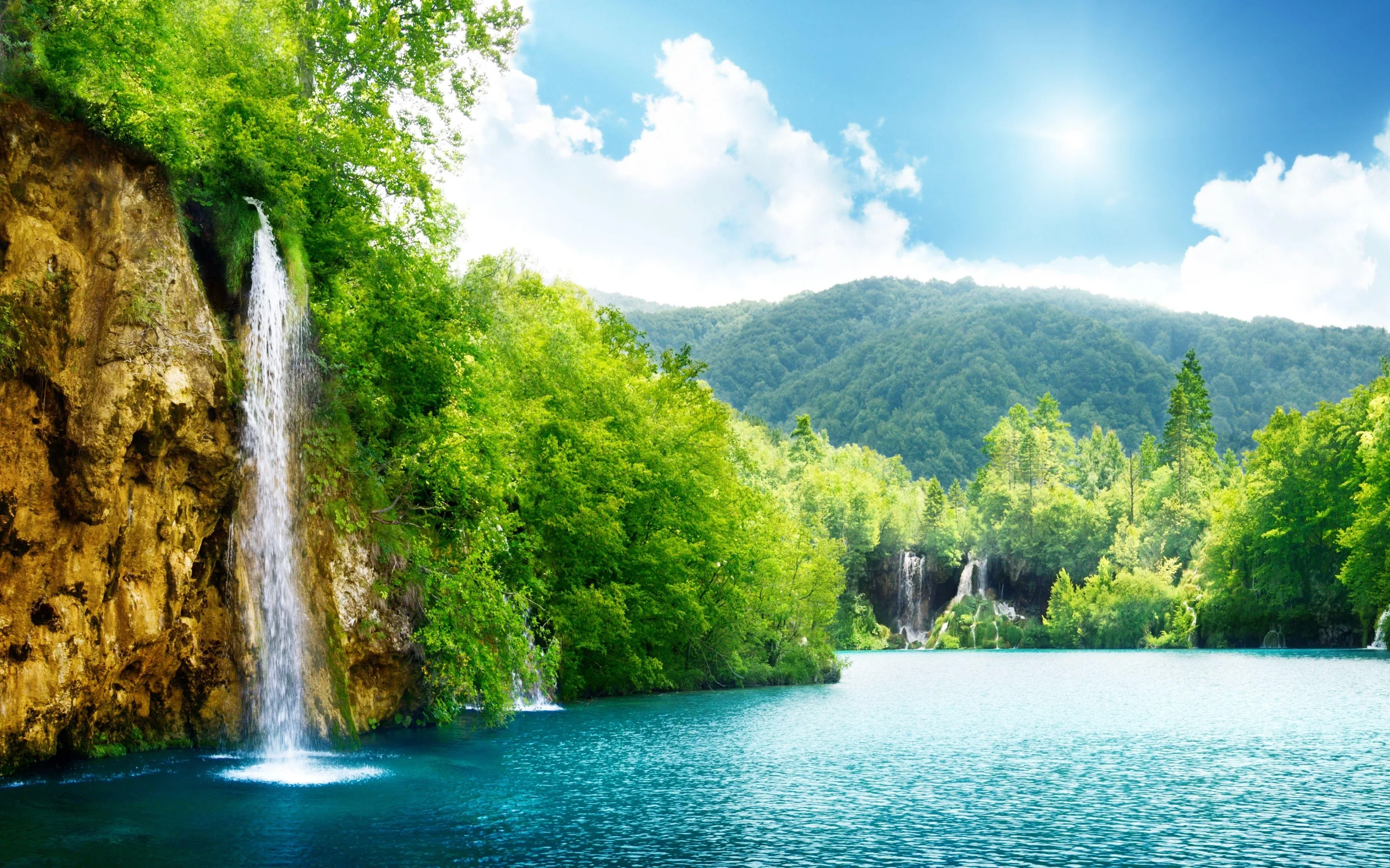 Water sheets down a rust-stained cliff at left while the sun flares in the top corner, throwing a bright track across the turquoise lake.