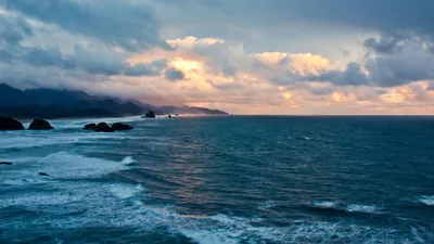 Rows of teal swell push toward dark headlands and sea stacks, a patch of sunset gold breaking through the clouds just right of centre.