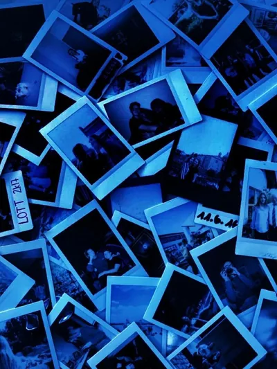 Instant photo prints lie scattered in a loose pile under deep blue light, their white borders glowing while the gaps between them drop to black.