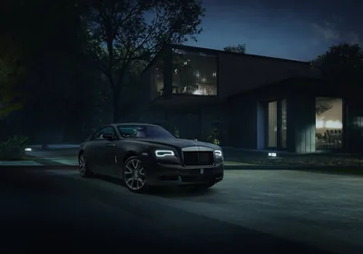 A long black coupe on the gravel drive of a lit glass-and-timber house, the tail lamps glowing and the trees above lost in blue darkness.