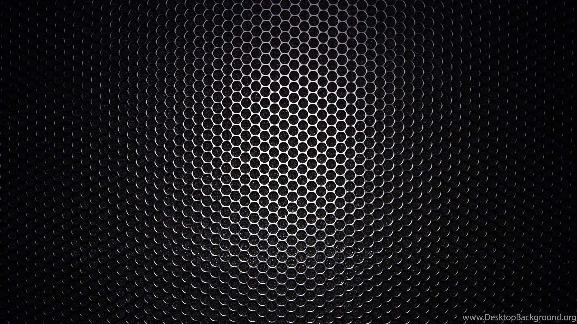 A wide field of small round holes in dark metal, lit from behind at the centre and falling away to black on both sides.