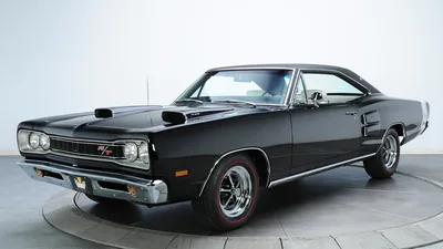 A triple black Coronet R/T with a twin-scooped hood stands on a pale studio floor, red-striped tyres and chrome bumpers picked out by even overhead light.
