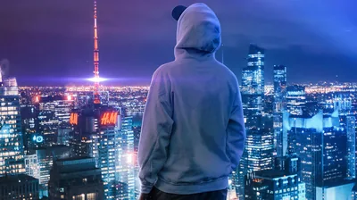 Shot from behind in a pale hoodie, he looks out over a violet skyline with a lit broadcast tower rising on the left.