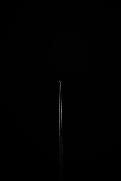 A needle-fine vertical streak rises through a tall black frame, a barely visible round shape hanging just behind its tip.