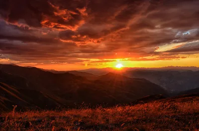 The sun drops between distant ridges under low red-brown clouds, a grassy slope in the foreground catching the final warm rays.