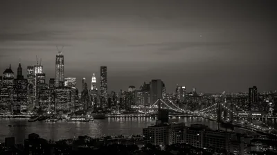 The full skyline and a suspension bridge sit under a warm grey sky, lights spread thin along the waterfront.