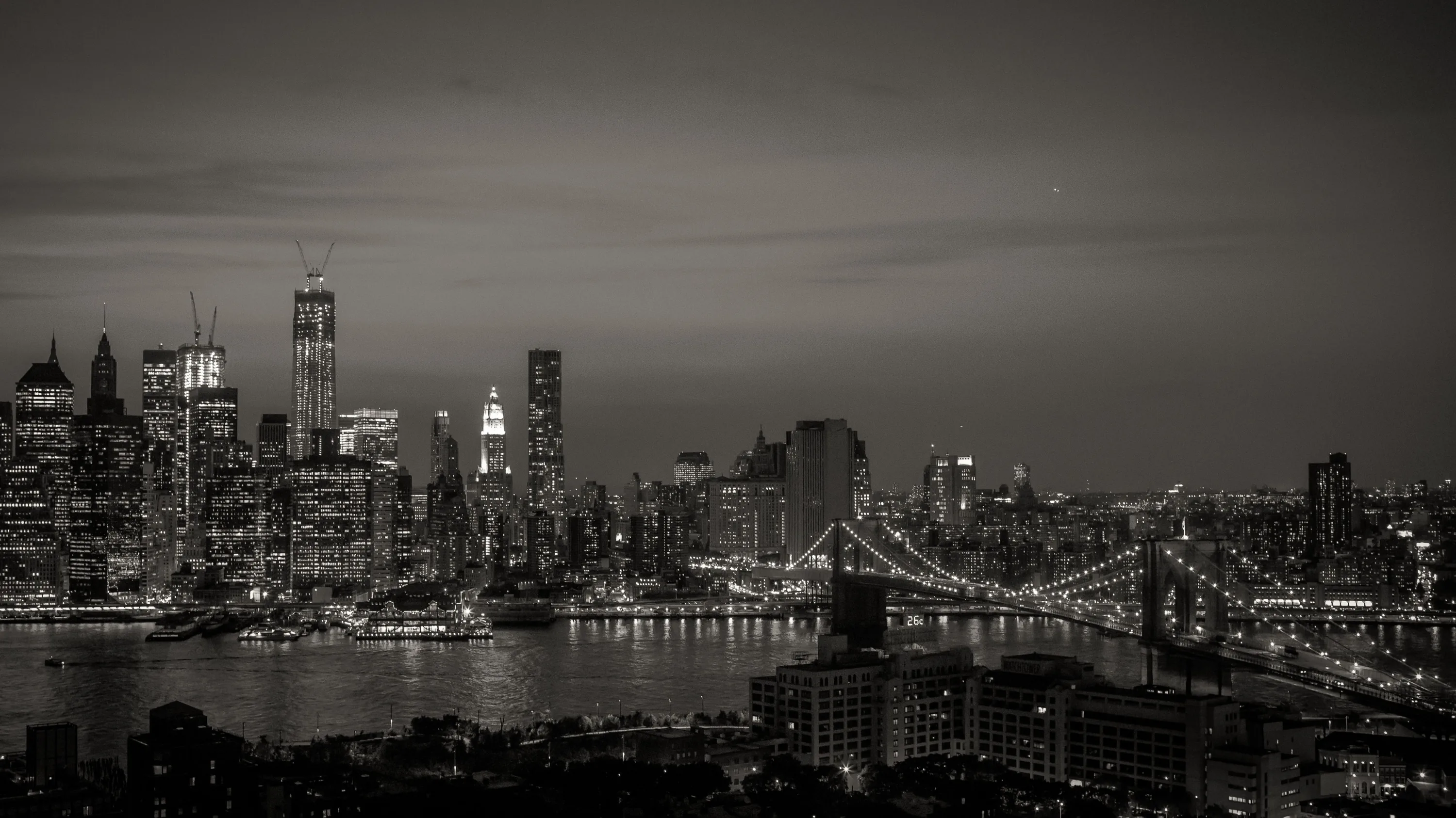 The full skyline and a suspension bridge sit under a warm grey sky, lights spread thin along the waterfront.
