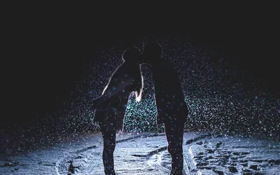 Two figures lean into a kiss on a snowy path at night, backlight turning every falling flake into a bright speck around them.