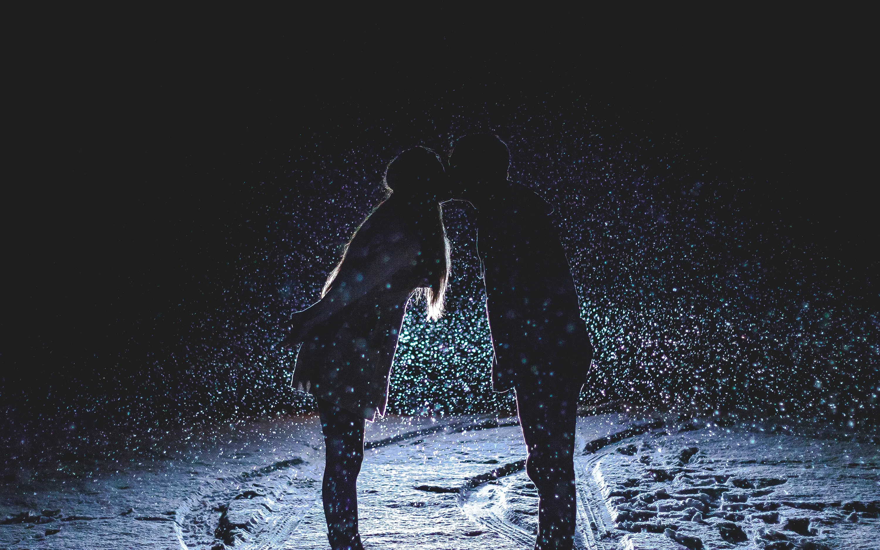 Two figures lean into a kiss on a snowy path at night, backlight turning every falling flake into a bright speck around them.