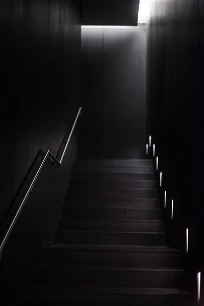 A wooden staircase climbs toward a single lit landing, small floor lights marking the right-hand edge of each tread.