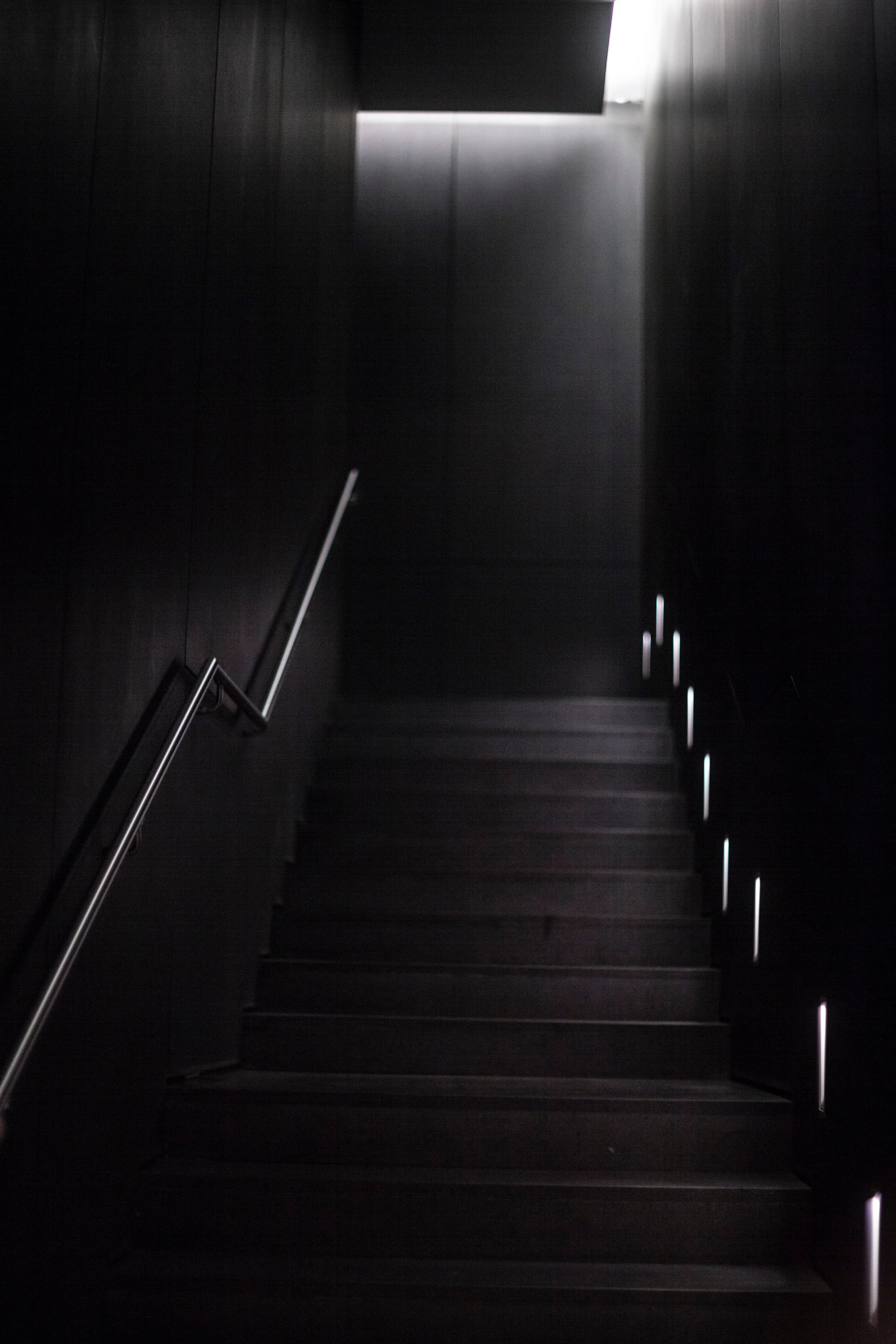 A wooden staircase climbs toward a single lit landing, small floor lights marking the right-hand edge of each tread.