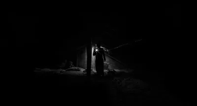 A shadowed figure stands in a narrow lit opening, the rest of the scene sunk into deep black with only a hint of ground.