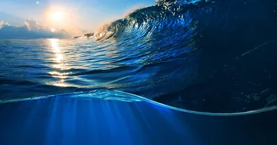 A deep blue wave curls at the surface while the frame dips below the waterline, sun rays fanning down through the glassy water beneath.