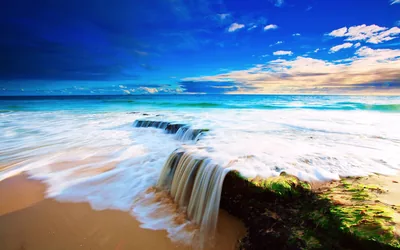 Backwash pours off a low rock shelf like a small waterfall, silky white water framed by wet sand and a saturated blue sky.