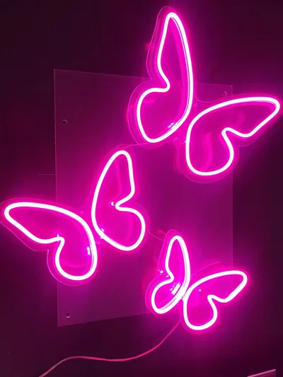 Three butterfly outlines in pink LED tube glow on a clear panel against a dark wall, the power cord trailing away at the bottom left.