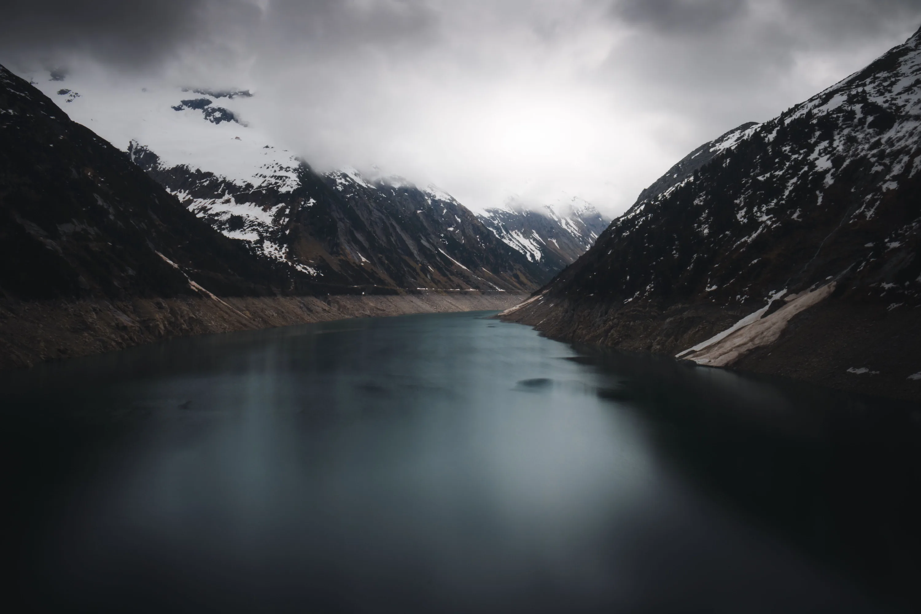Still grey water runs between snow-streaked slopes with low cloud sitting on the peaks at the far end.