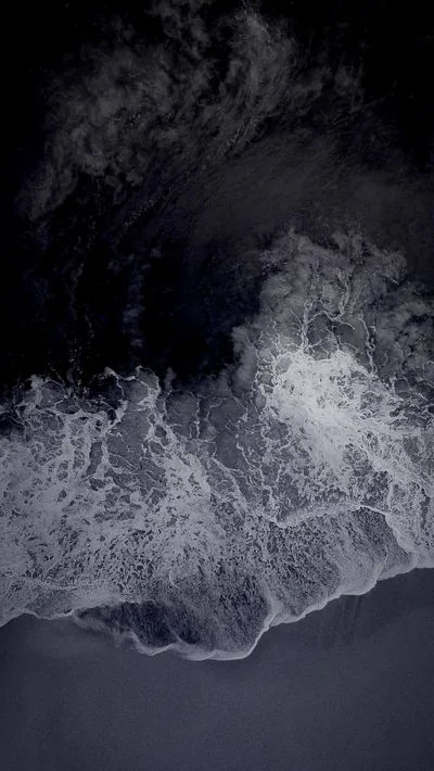 Shot straight down at the shoreline, white surf tears across dark volcanic sand while the deep water at the top stays near black.