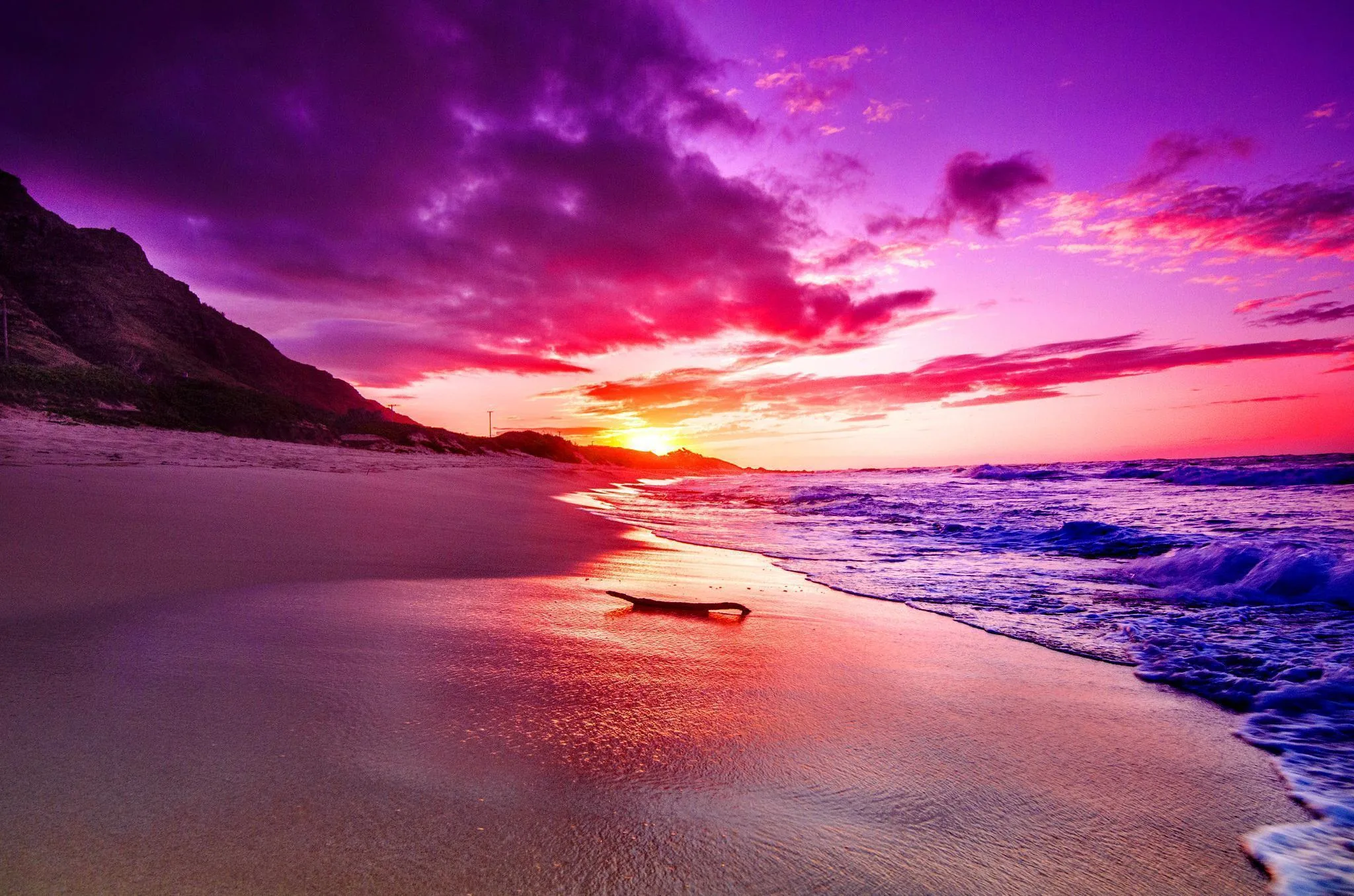 Purple and pink clouds roll over a dark headland while blue surf follows the curve of the sand, a piece of driftwood resting mid-beach.