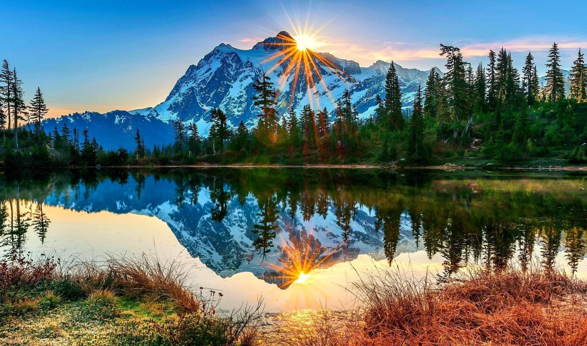 The sun breaks over a glaciated summit ringed by conifers, the lake repeating peak, trees and flare across the lower half of the frame.