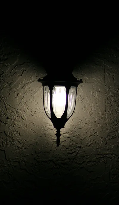 An old lantern-style lamp throws twin shadow wings up a rough stucco wall, the bottom third falling dark enough for icons.