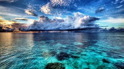 A massive cumulus bank rolls above glassy turquoise water, dark coral heads showing through the shallows along the bottom of the frame.