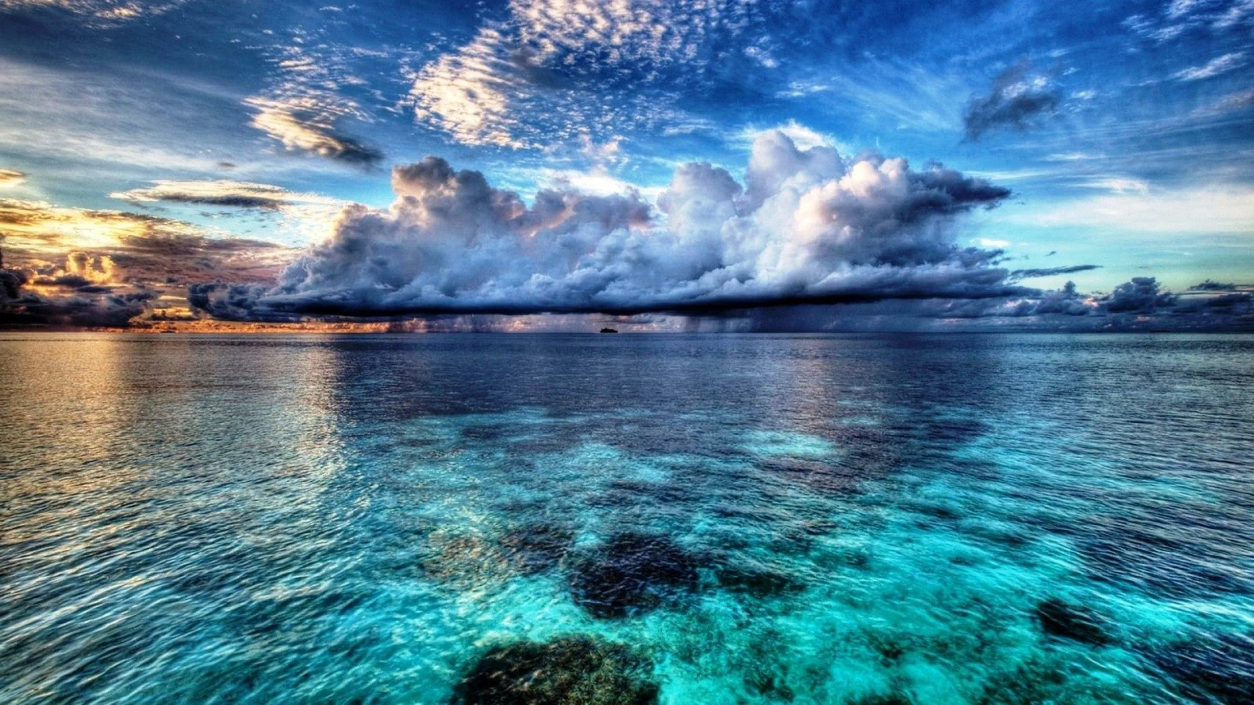 A massive cumulus bank rolls above glassy turquoise water, dark coral heads showing through the shallows along the bottom of the frame.