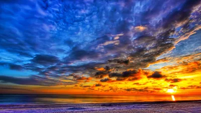 Heavy blue cloud presses down on a burning orange horizon in a high-contrast HDR beach scene, the sun touching the sea at lower right.