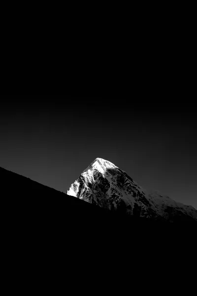 A snow-covered summit catches sunlight while the ridge in front of it drops away as a solid black wedge across the lower frame.