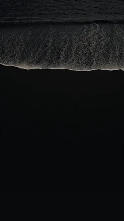 An aerial view where a thin white wave meets black volcanic sand, the lower half almost pure black.