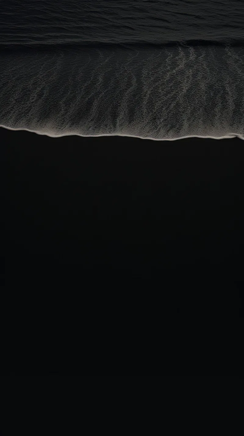 An aerial view where a thin white wave meets black volcanic sand, the lower half almost pure black.