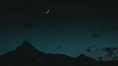 A thin crescent hangs high left of centre in a teal-black sky, the jagged mountain silhouette holding down the lower left corner.