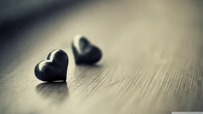 A pair of small dark hearts rest on bare wood, the front one sharp and the second blurred into a desaturated background.