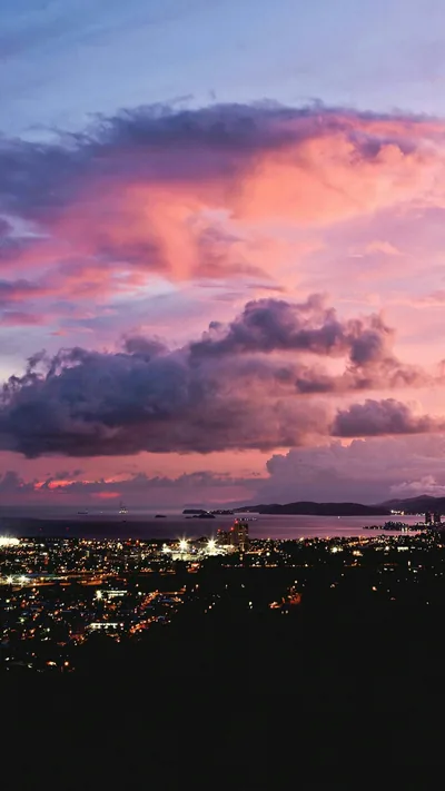 Pink and violet storm clouds stack above a coastal city at dusk, its street lights forming a glowing band across the lower third.