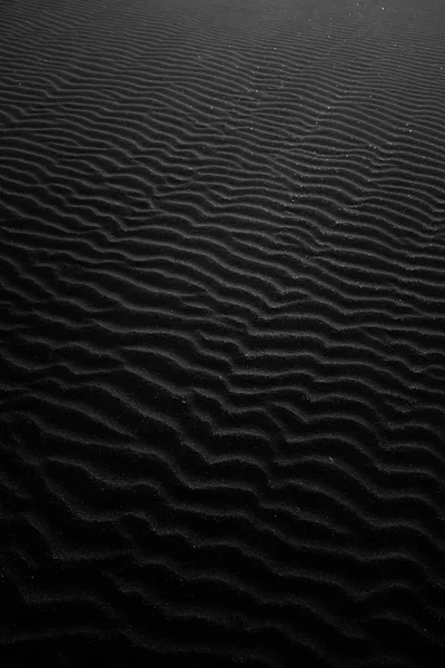 Rows of wind-cut ripples run over dark volcanic sand in a tall frame, the grain sharpest along the bottom and softening toward the top.