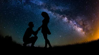 A kneeling figure holds a partner's hand on a dark hilltop while the Milky Way arcs overhead and a warm glow sits on the horizon.