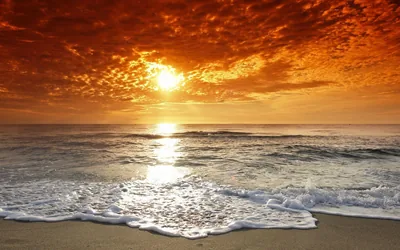 A ceiling of red-orange cloud burns overhead while foam slides up pale sand, the sun's reflection dropping a bright column down the middle of the frame.