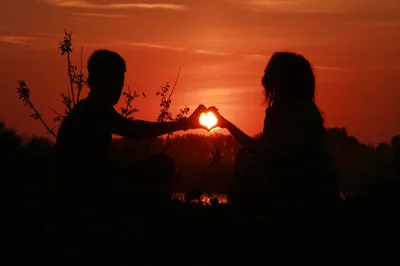A man and a woman face each other at dusk and touch fingertips so the setting sun sits inside the heart their hands make, everything else in dark red silhouette.