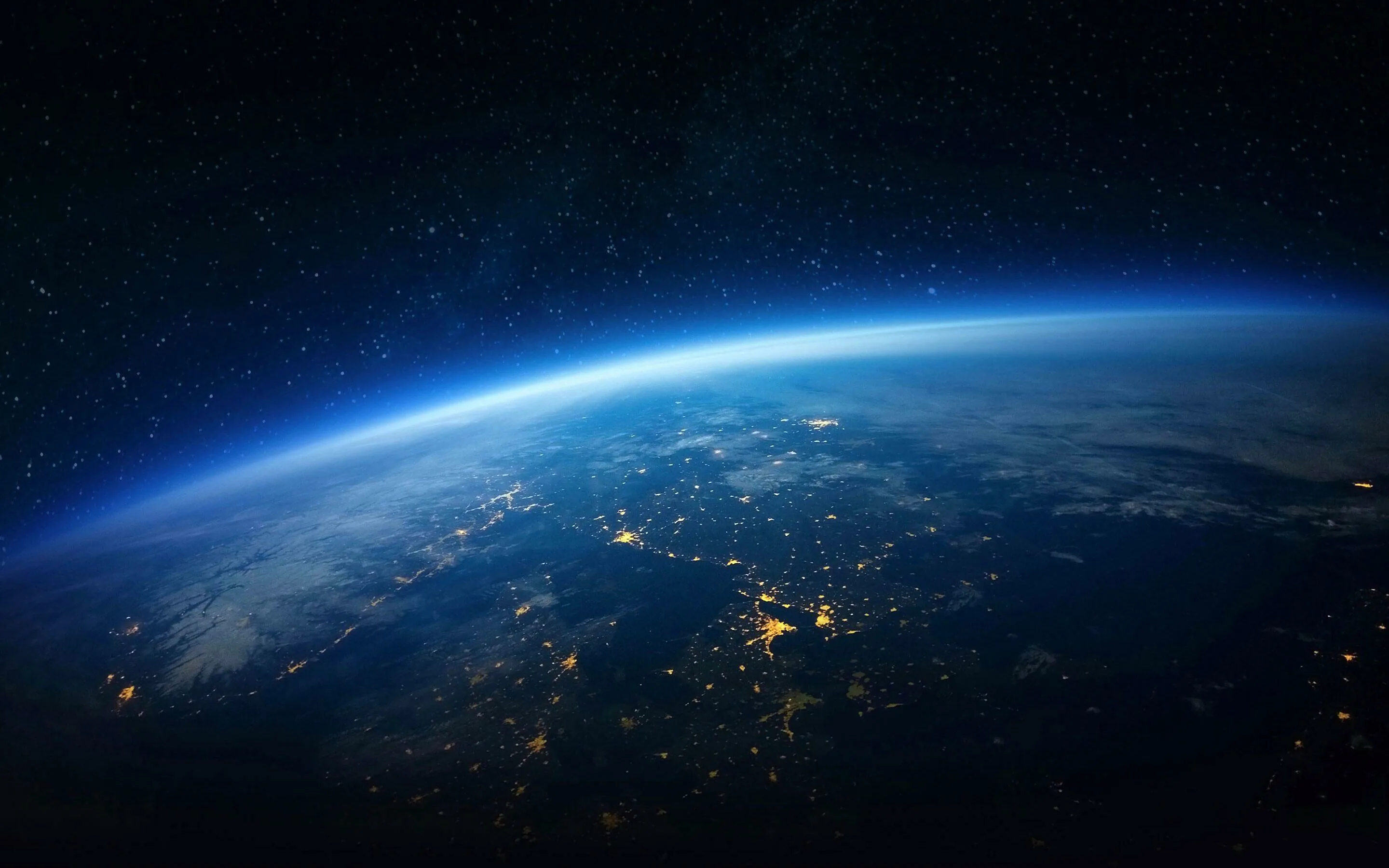 Earth's night side curves across the frame under a faint blue atmospheric arc, scattered city lights in the centre leaving both upper corners fully dark.
