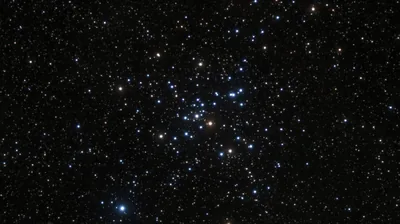 Blue and white cluster stars gather at the centre of this telescope image, one amber giant among them, thinning out toward black corners.