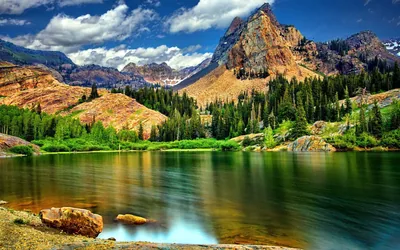 Iron-stained summits climb behind a pine shoreline while long exposure flattens the lake to glass, the darkest water sitting in the lower left corner.