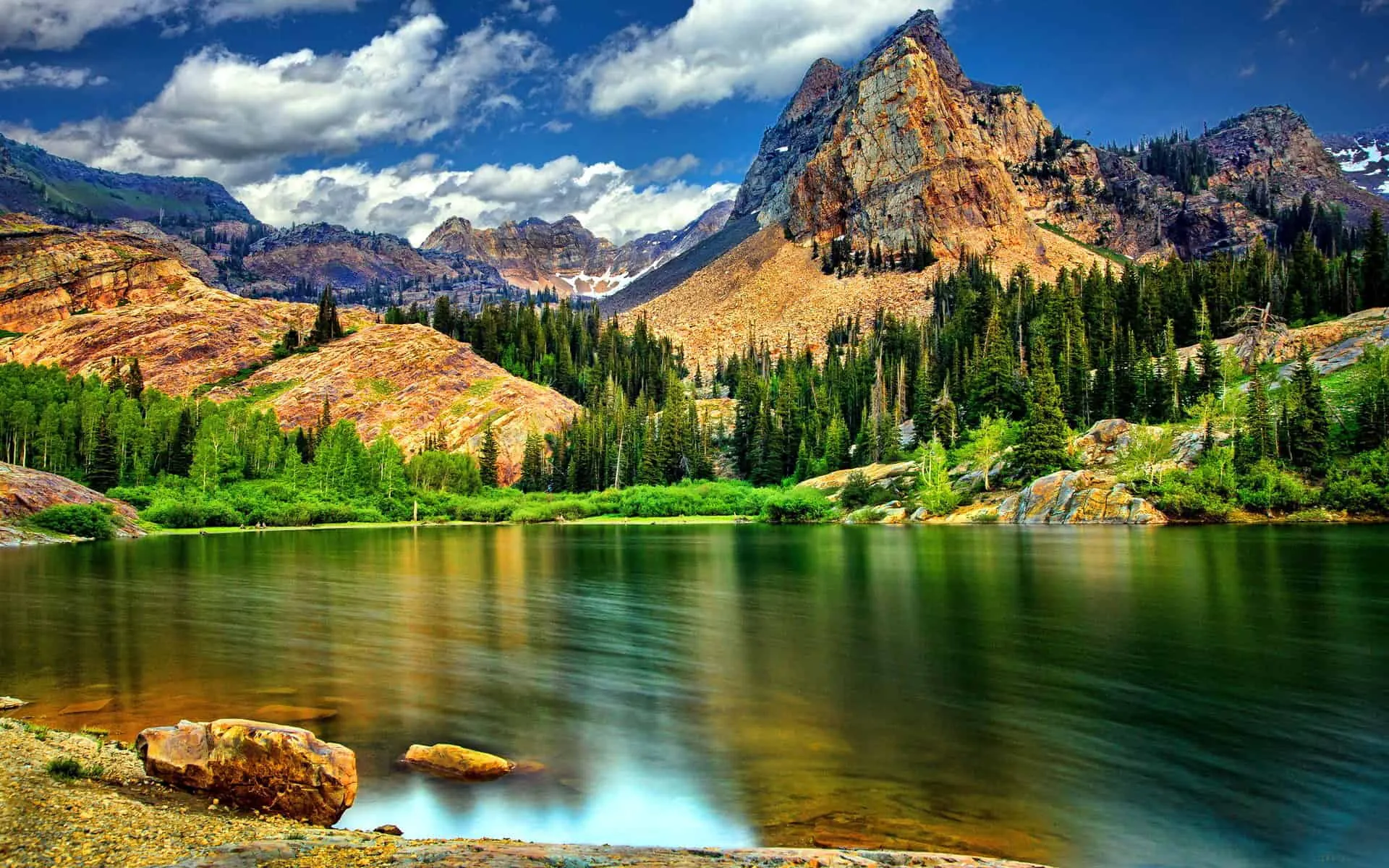 Iron-stained summits climb behind a pine shoreline while long exposure flattens the lake to glass, the darkest water sitting in the lower left corner.