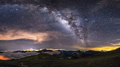 The Milky Way arcs over a dark mountain ridge in a stitched panorama, orange town glow leaking along the horizon at both ends.