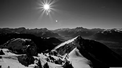 A hard starburst sun sits above a run of snow-covered ridges, the valleys below dropping into deep shadow.