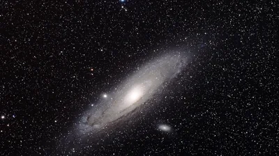 A large tilted galaxy sits left of centre in warm greyscale with two small companion galaxies nearby and stars spread thinly around it.
